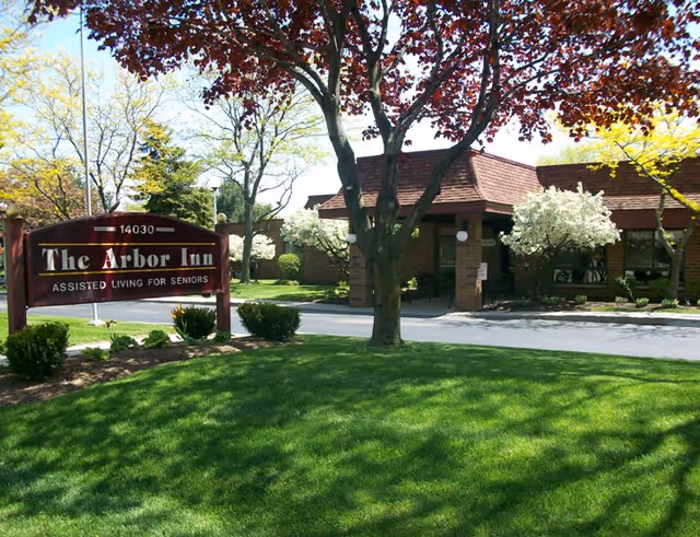 Exterior view of The Arbor Inn assisted living facility for seniors, showing a brick building with a brown roof, surrounded by green grass, trees with red and white blossoms, and a sign with the facility's name and address.