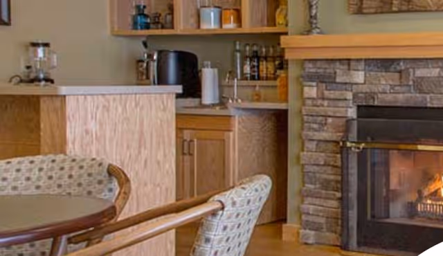 Cozy interior space featuring a round table with patterned chairs, a wooden kitchen counter with cabinets, and a stone fireplace with a fire burning inside.