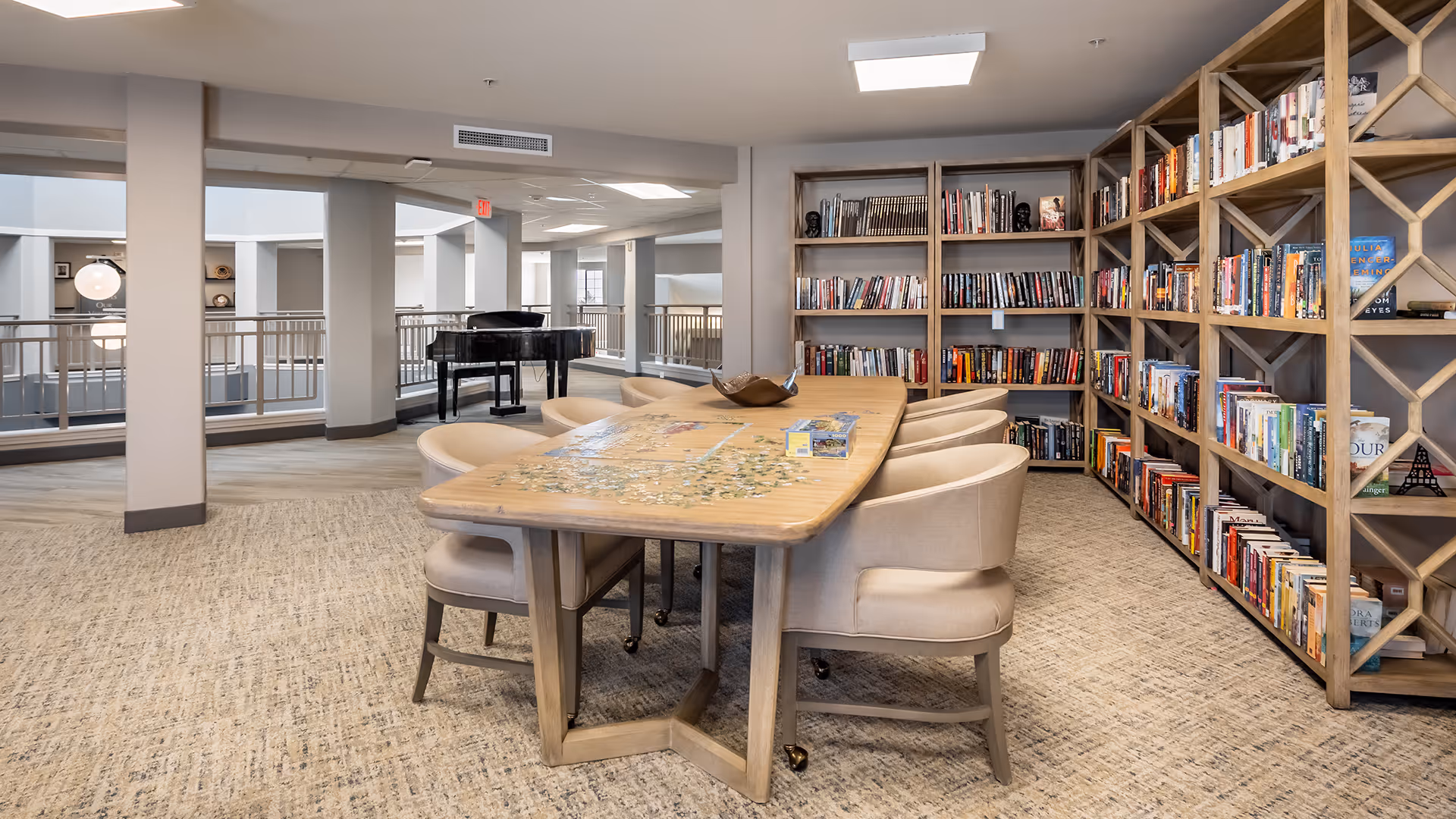 A cozy reading and activity room with a large wooden table surrounded by beige upholstered chairs. The table has a puzzle in progress on it. Behind the table, there are wooden bookshelves filled with books. In the background, a black grand piano is visible near a railing overlooking an open area with columns and windows.