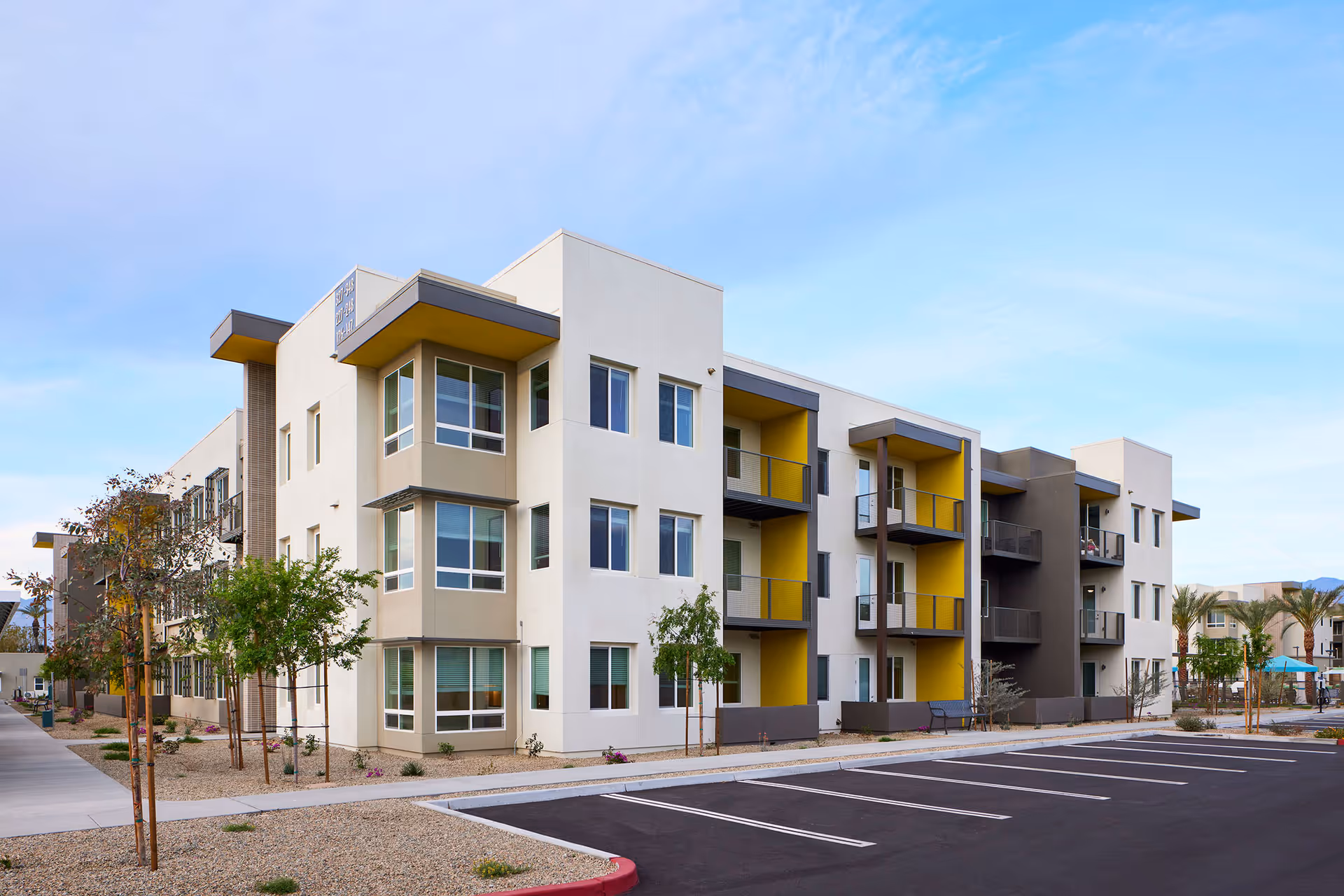 Modern three-story apartment-style building with balconies, yellow accent walls, landscaping and an empty parking lot.