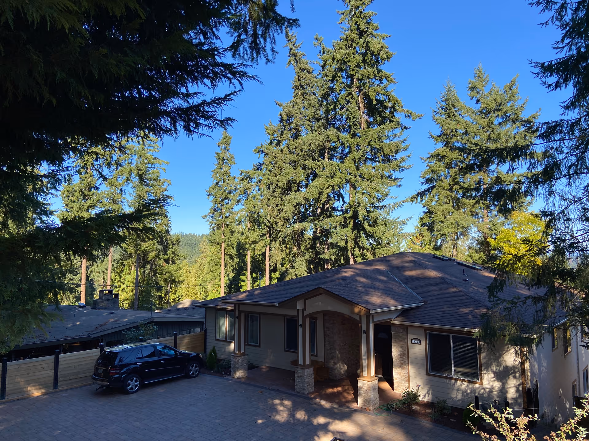 A single-story residential building surrounded by tall evergreen trees under a clear blue sky. There is a paved driveway with a black SUV parked on the left side. The building has a covered entrance with stone pillars and beige siding.