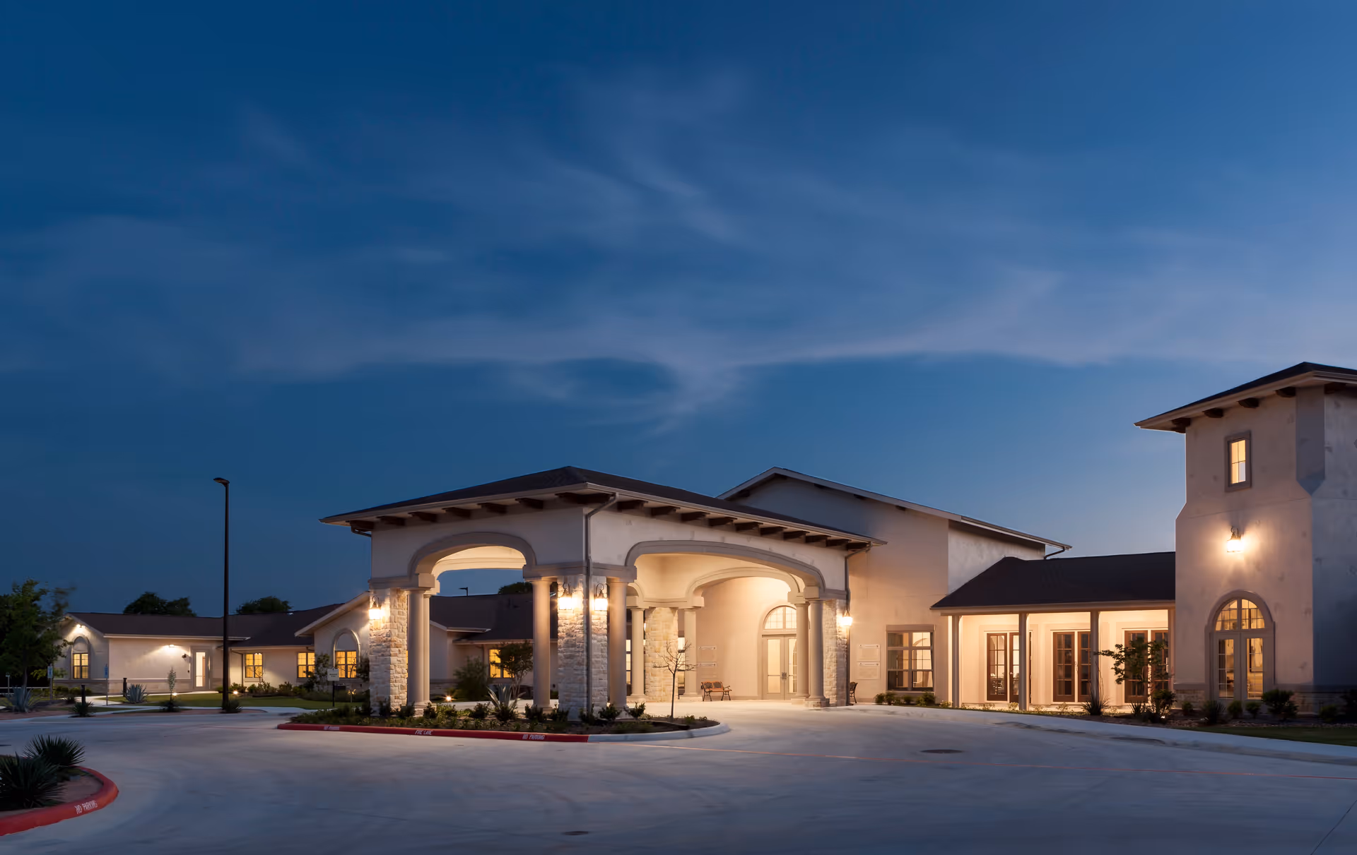 Exterior view of Franklin Park Boerne senior living facility at dusk, showing a well-lit entrance with columns and arched doorways, surrounded by a paved driveway and landscaped greenery under a clear evening sky.