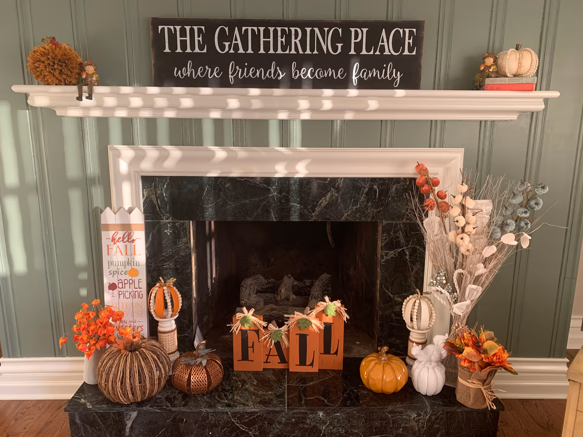 Decorated fireplace with a mantel sign reading 'The Gathering Place' and autumn pumpkins and floral arrangements.