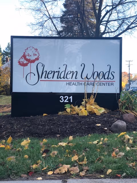 Outdoor sign for Sheriden Woods Health Care Center with the number 321 displayed below the name. The sign is surrounded by grass, fallen leaves, and some plants, with trees and houses visible in the background.