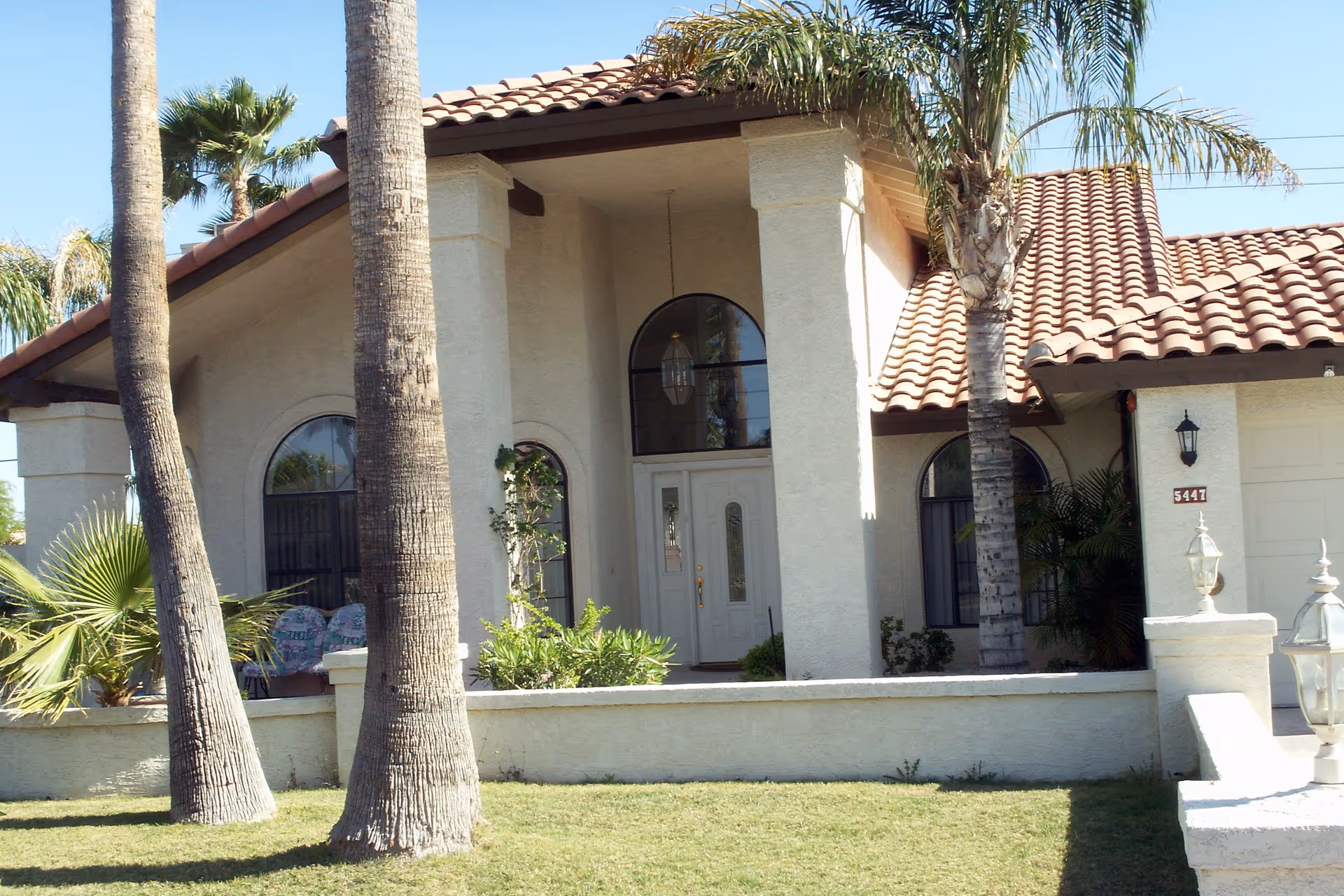Front exterior view of a single-story house with a tiled roof, arched windows, a white front door with glass panels, palm trees, and a small garden area in front.