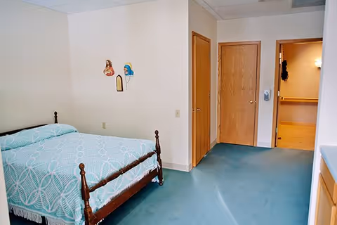 A simple bedroom with a wooden bed frame and a light blue patterned bedspread. The room has light-colored walls with two small religious wall decorations. There are two wooden doors and an open doorway leading to a hallway with wooden flooring. A hand sanitizer dispenser is mounted on the wall near the doors.
