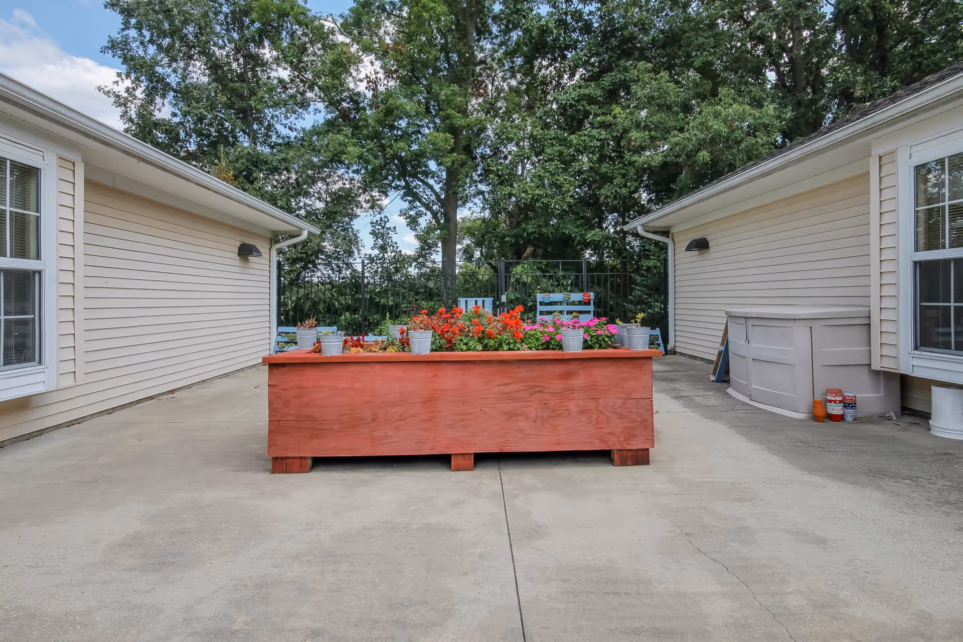 Outdoor patio area between two beige buildings with a large red wooden planter box filled with colorful flowers in the center. Trees and a blue sky with clouds are visible in the background.