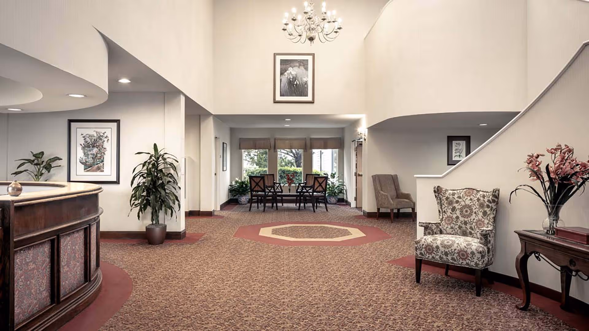 A spacious and well-lit senior living facility lobby with patterned carpet flooring, a curved wooden reception desk on the left, several upholstered chairs, a small table with a vase of flowers on the right, potted plants, framed artwork on the walls, and a chandelier hanging from the ceiling.