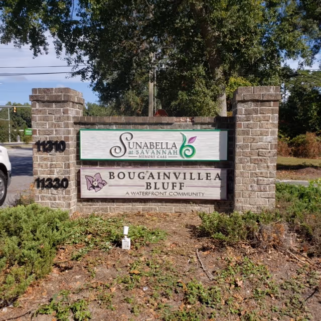A brick signpost with two signs mounted on it. The top sign reads 'Sunabella at Savannah Memory Care' with a green and purple leaf logo. The bottom sign reads 'Bougainvillea Bluff A Waterfront Community' with a purple flower logo. The signpost is surrounded by greenery and trees in the background.
