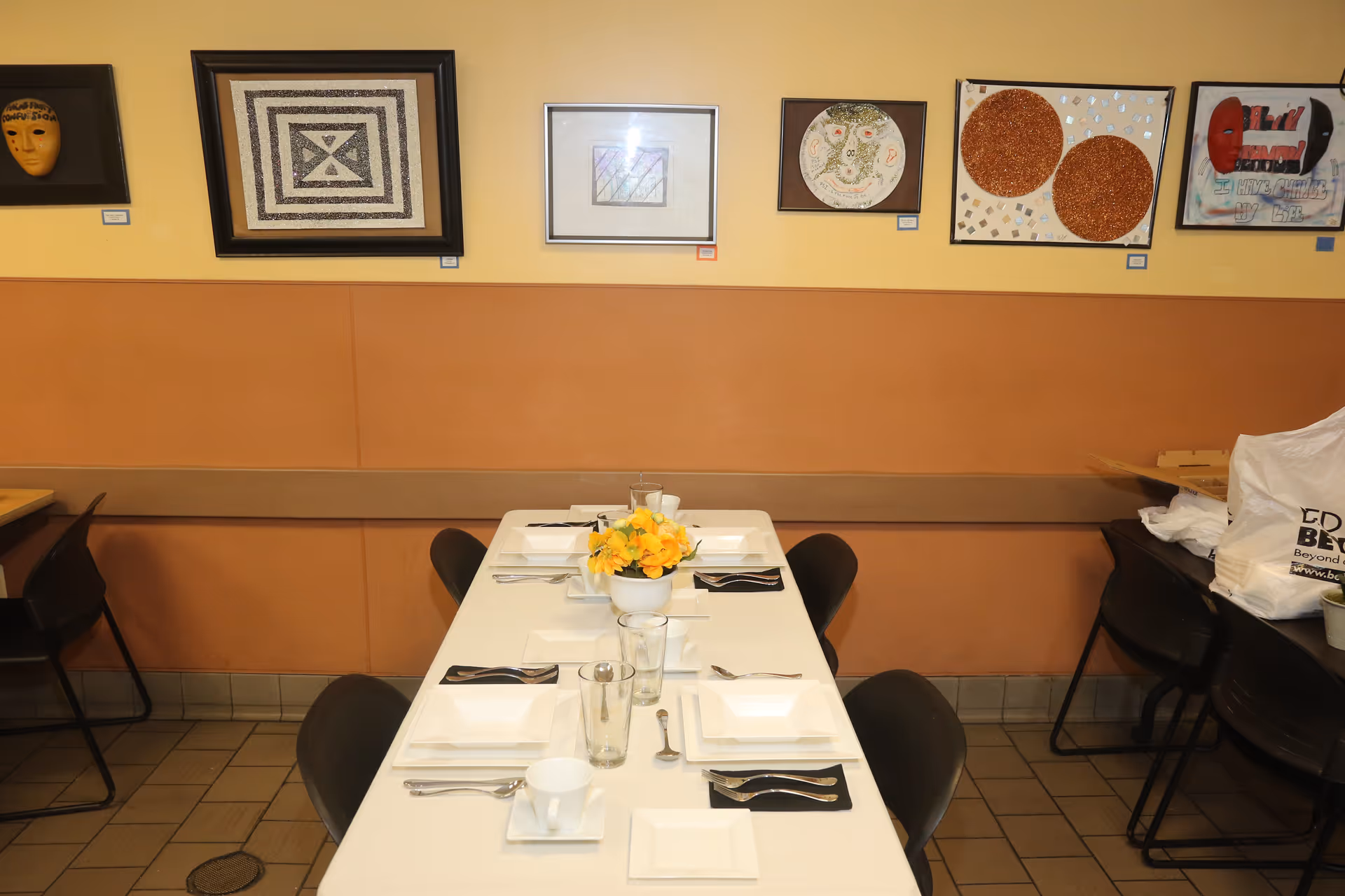 A dining area with a rectangular table set for six people with white square plates, cups, glasses, and silverware on black napkins. A small flower arrangement with yellow flowers is in the center of the table. The walls are painted in two tones of beige and decorated with various framed artworks. Black chairs surround the table, and there are additional chairs and tables visible on the sides.