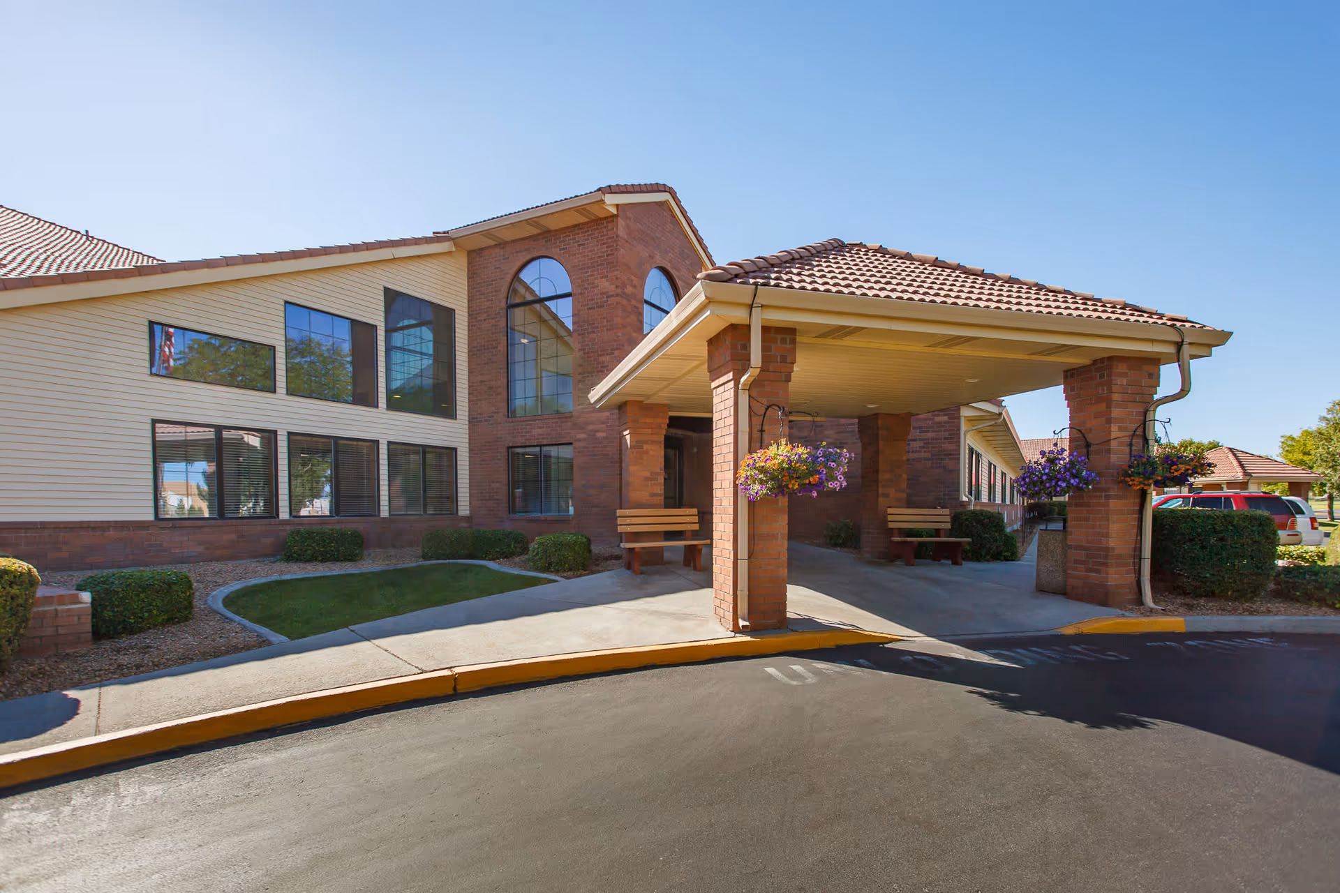 Exterior view of Bridgeview Estates senior living facility showing a covered entrance with brick pillars, hanging flower baskets, benches, and a well-maintained lawn under a clear blue sky.