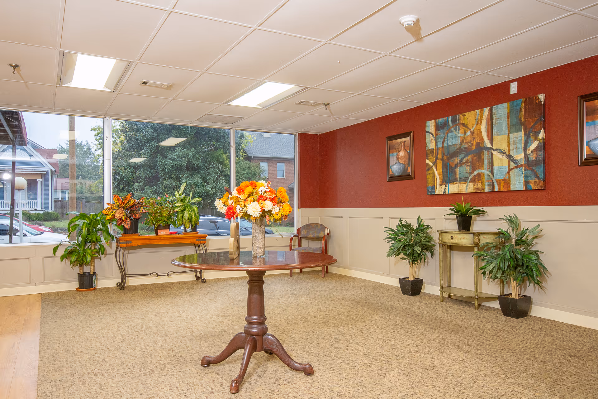 Bright common room with a round wooden table topped by a vase of flowers, potted plants, artwork on red walls, and large windows.