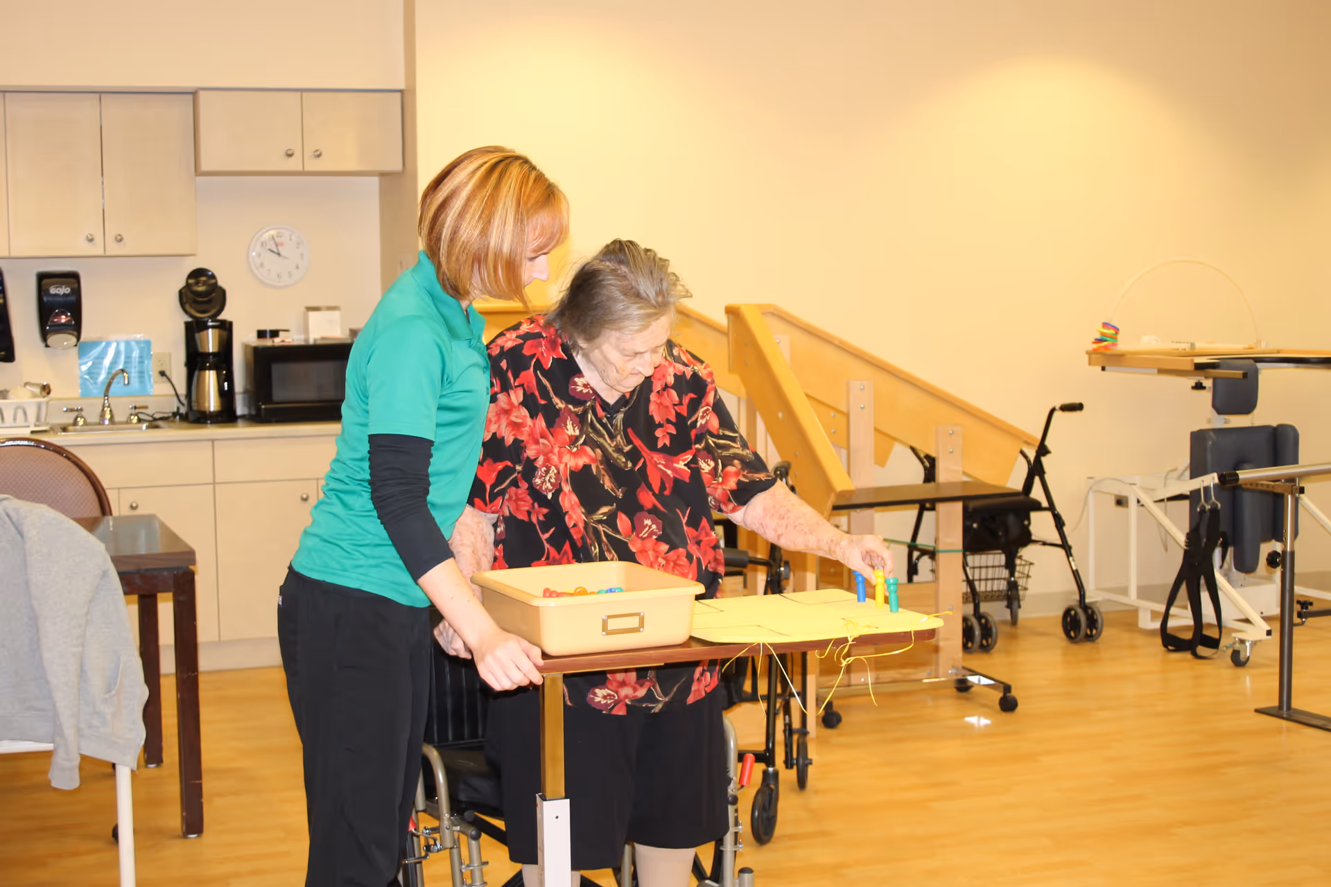 A caregiver assists an elderly woman with a tabletop activity in a bright interior therapy/recreation room with mobility aids and a kitchenette in the background.