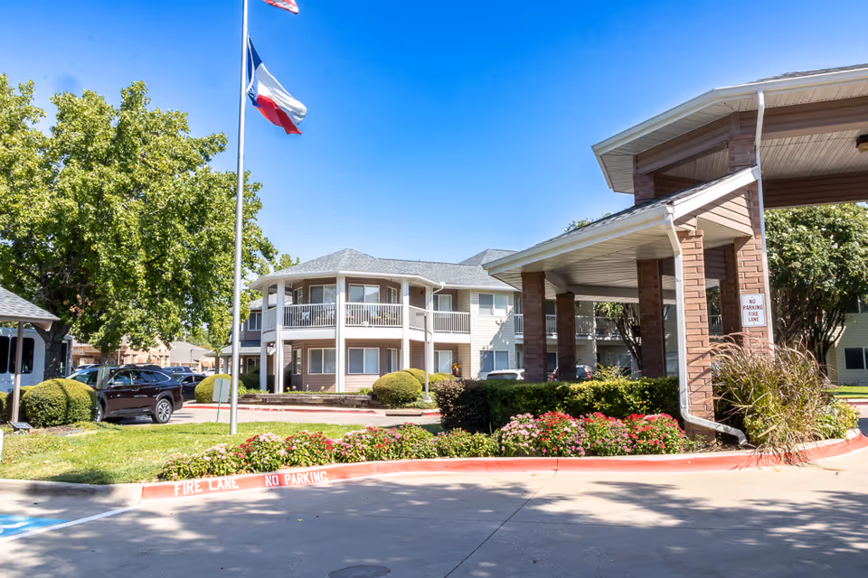 Exterior view of Asher Point Independent Living of Arlington showing a two-story building with balconies, a covered entrance supported by brick columns, a flagpole with the Texas state flag, well-maintained landscaping with bushes and flowers, and a clear blue sky.