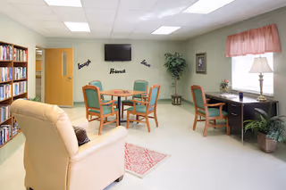 A cozy common area with a beige armchair, a round table surrounded by four green chairs, a bookshelf filled with books, a wall-mounted TV, and a desk with two chairs near a window with pink curtains. The walls are light-colored and decorated with words 'laugh', 'friends', and 'love', along with a framed picture and a potted plant.