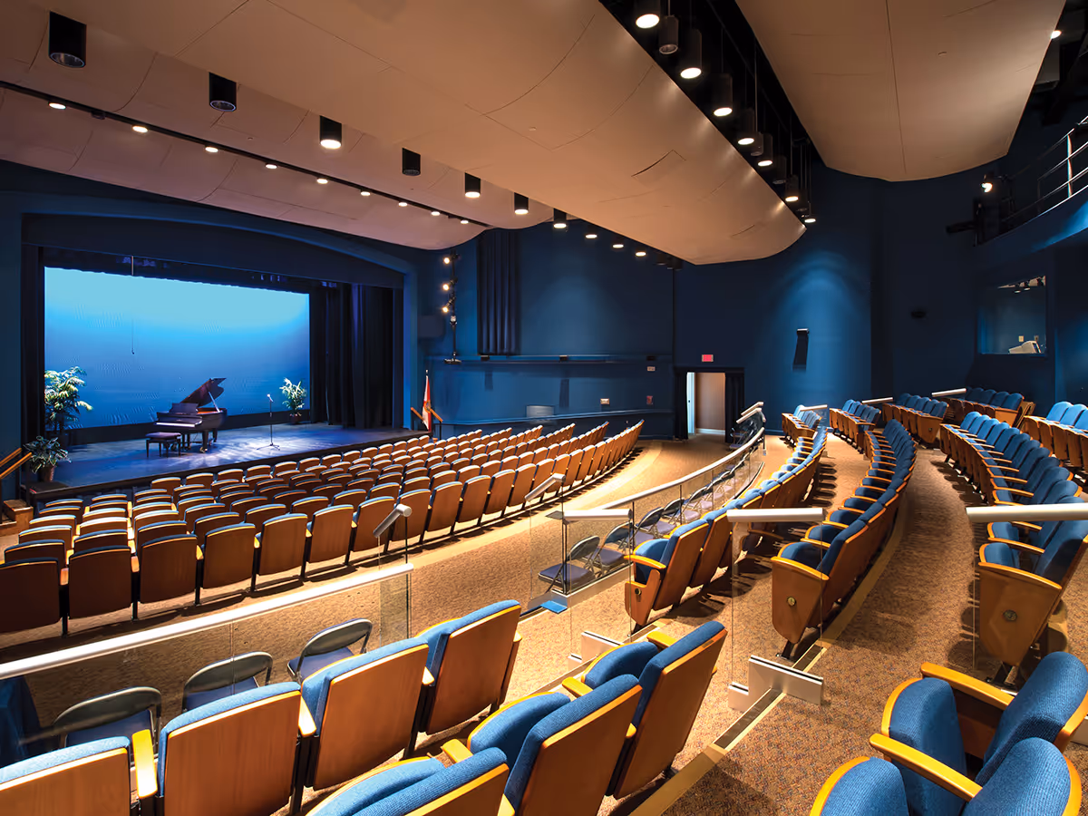 Interior view of an auditorium with rows of blue cushioned seats facing a stage with a grand piano and microphone stand, illuminated by ceiling lights.