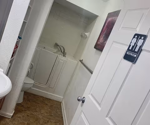 View into a small bathroom with a walk-in bathtub, a toilet, a sink, and a door with a restroom sign showing male and female icons. There is a handrail on the wall and a framed picture hanging above the bathtub.