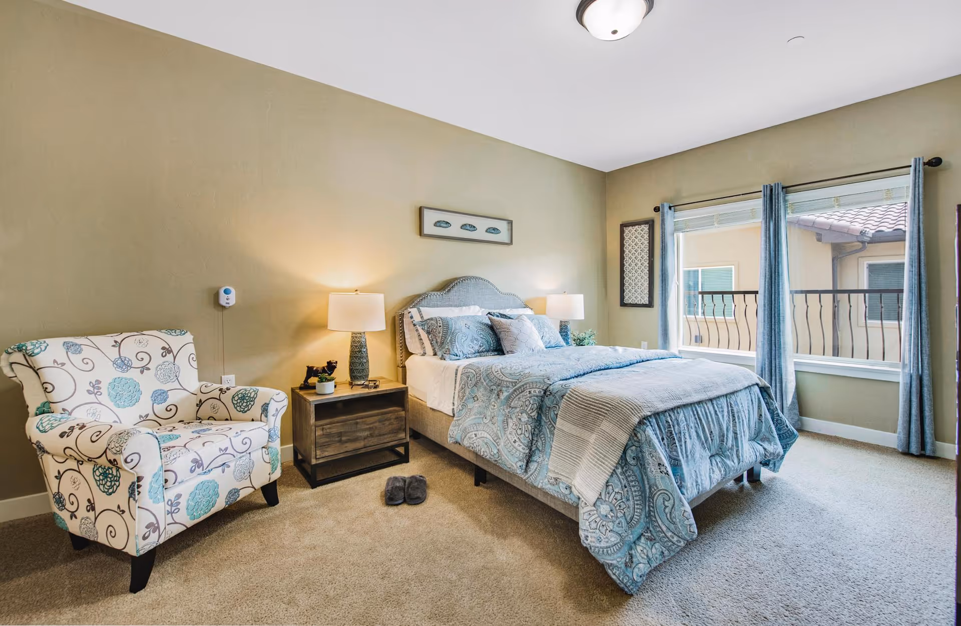 A cozy bedroom with a bed dressed in blue patterned bedding, a wooden nightstand with a lamp and small plant, a floral upholstered armchair, and large windows with blue curtains letting in natural light.