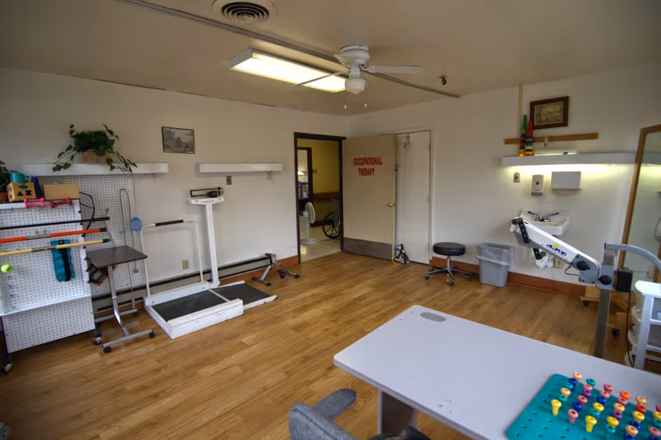 A therapy room with wooden flooring and white walls, containing various occupational therapy equipment including a scale, exercise tools, a stool, a sink, and a table with colorful pegboard. A door labeled 'Occupational Therapy' is visible, along with a wheelchair in the adjacent room.