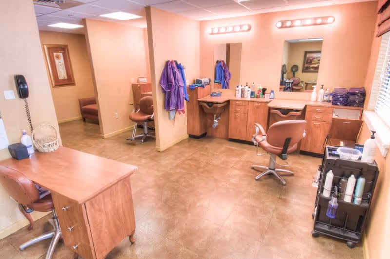 Interior view of a salon area in a retirement community with salon chairs, mirrors, wooden cabinets, hair care products, and hanging salon capes. The room has beige walls and tiled flooring with bright overhead lighting.