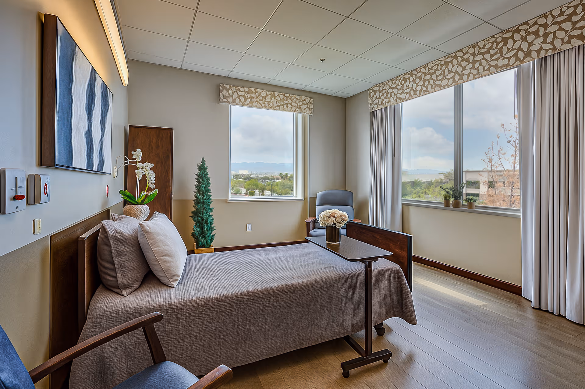Bright, neatly staged patient bedroom with a single bed, armchairs, a small overbed table with flowers, and large windows overlooking greenery.