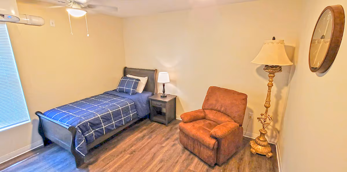 Small bedroom with a single bed, bedside table and lamp, a recliner and ornate floor lamp, and a wall clock.