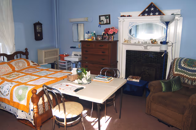 A cozy bedroom with a wooden bed covered by a colorful quilt, a small table with chairs, a brown armchair with a crocheted blanket, a wooden dresser with personal items and photos, a white fireplace with an oval mirror and a folded American flag displayed on top, and a small sink with toiletries in the corner.