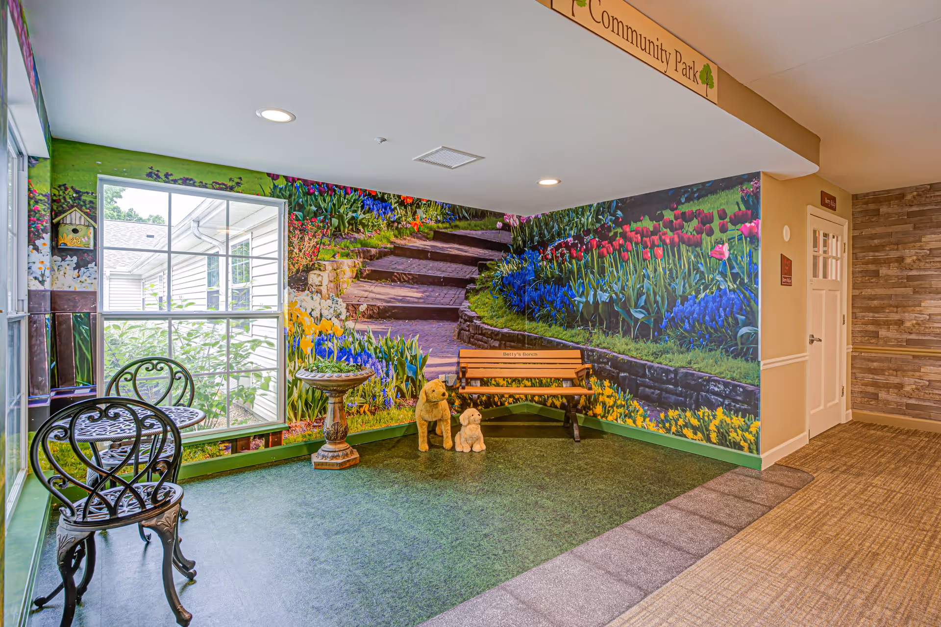 Indoor seating area with a small black metal table and two chairs near a window. The walls feature a large mural of a garden with colorful flowers, stone steps, and greenery. There is a wooden bench with two dog statues in front of it. The floor has a green carpet resembling grass and a beige carpeted hallway to the right.