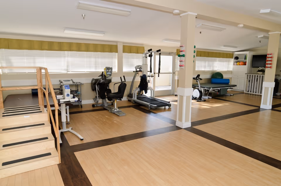 A spacious rehabilitation or physical therapy room with exercise equipment including a treadmill, stationary bike, parallel bars, and therapy tables. The room has large windows with blinds, wooden flooring with dark inlays, and light-colored walls with columns. Small dumbbells are mounted on the columns, and there is a TV mounted on the wall in the background.