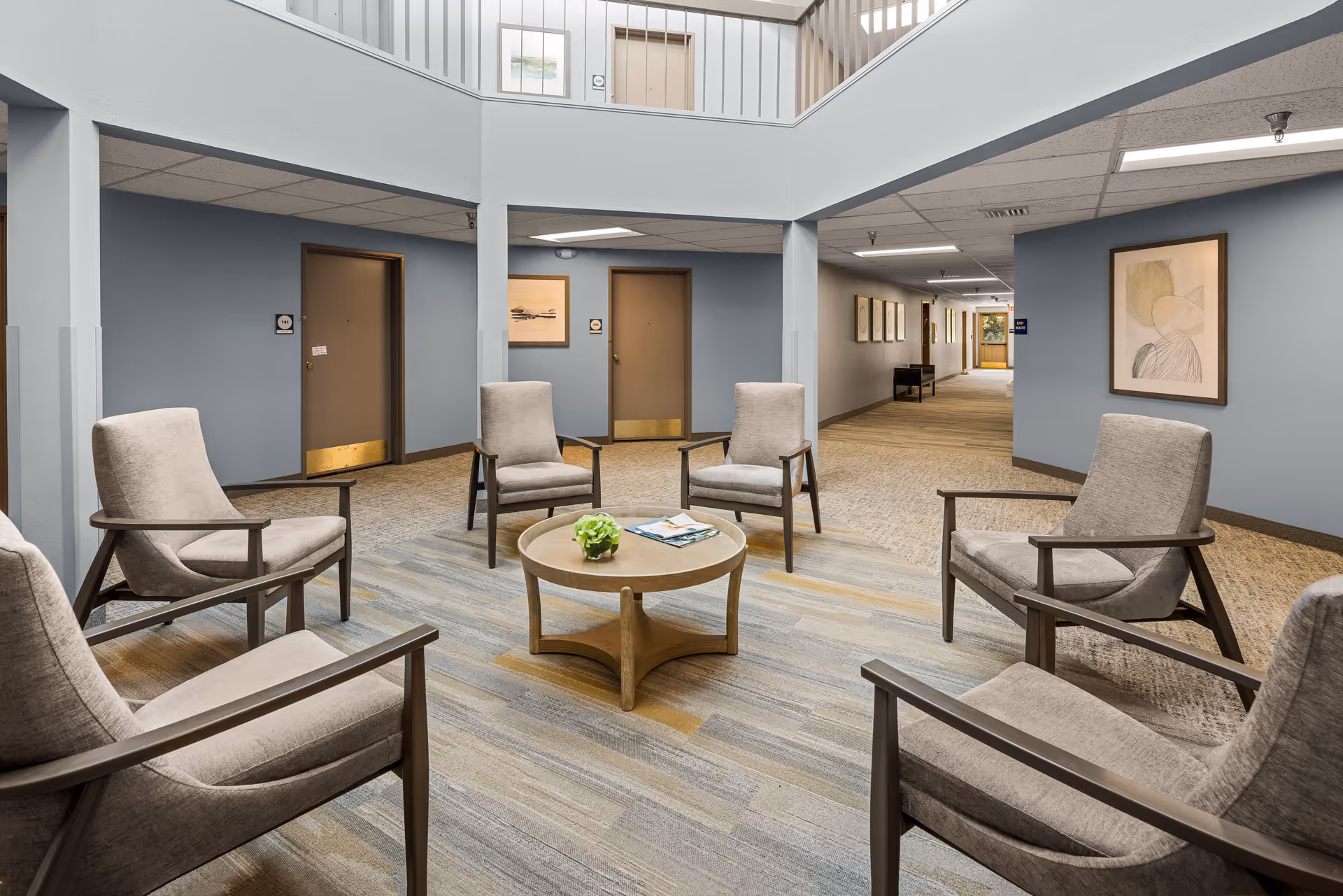 A seating area in a senior living facility with six beige armchairs arranged in a circle around a round wooden coffee table with a small green plant and magazines. The space has blue-gray walls, carpeted floors, and doors leading to other rooms. There is a hallway visible in the background with framed artwork on the walls.