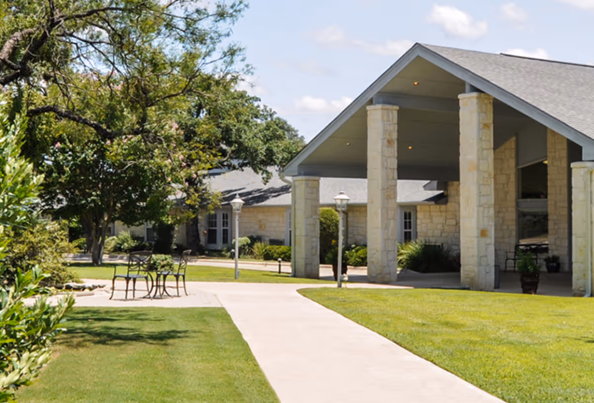 Outdoor view of a senior living facility with a stone building featuring a covered entrance supported by stone pillars. There is a paved walkway leading to the entrance, surrounded by well-maintained green lawns, trees, and shrubs. A small patio area with a table and chairs is visible on the left side under the shade of a tree.