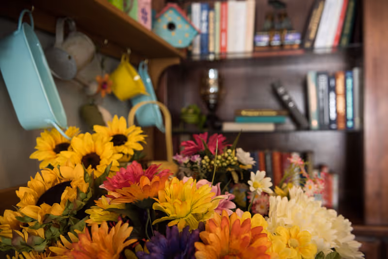 A close-up view of a colorful bouquet of artificial flowers including sunflowers and daisies in front of a wooden bookshelf filled with books and decorative items such as small birdhouses and hanging pastel-colored metal containers.