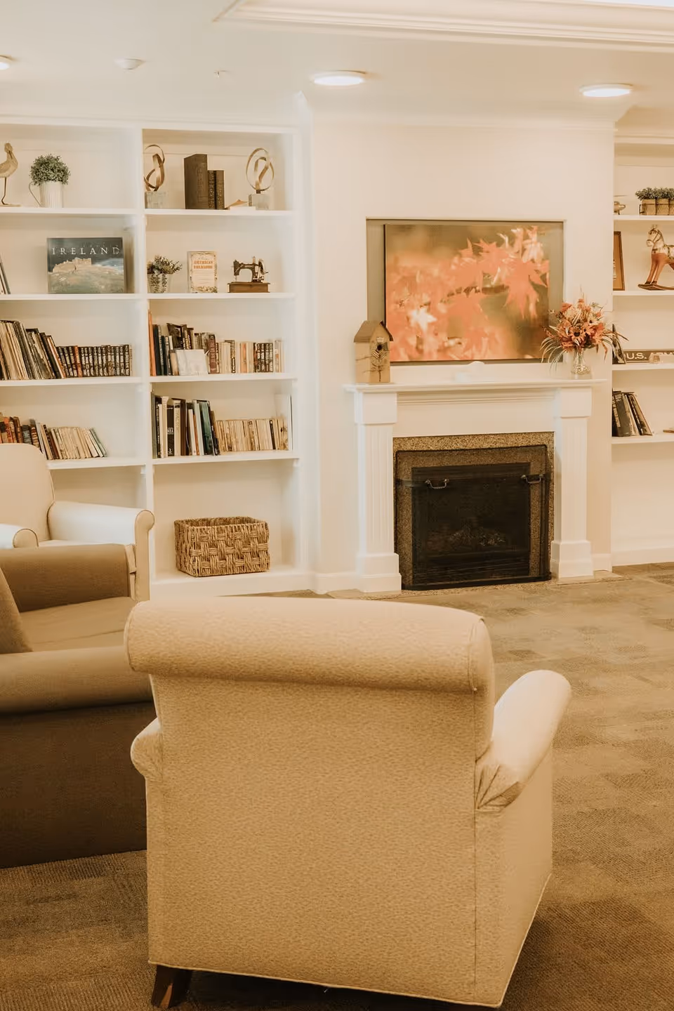 A cozy living room area with beige and brown armchairs facing a white fireplace. Above the fireplace is a framed picture of autumn leaves. Built-in white bookshelves flank the fireplace, filled with books, decorative items, and plants. The room has a warm and inviting atmosphere with soft lighting.