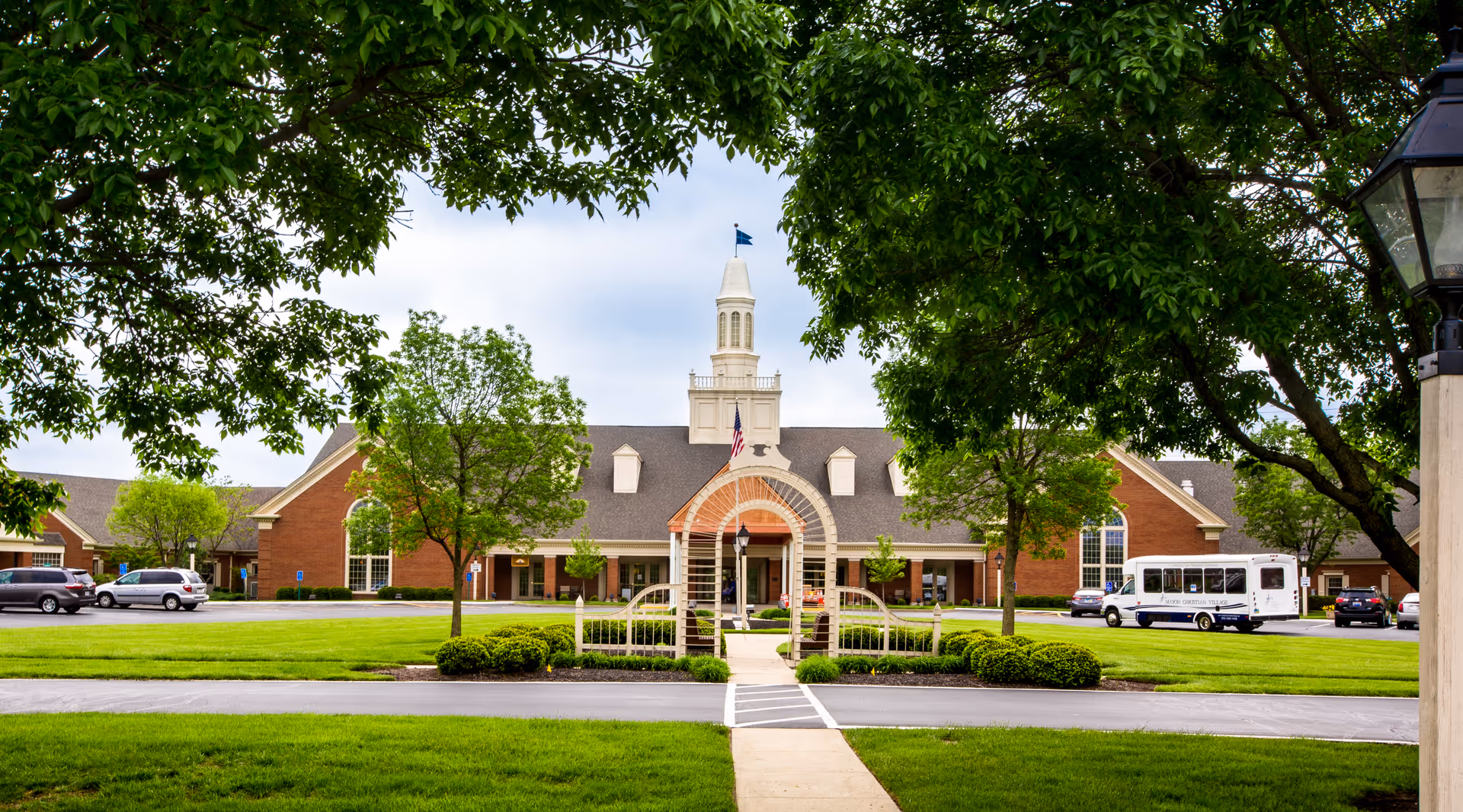 Front exterior view of The Christian Village at Mason building with a central archway entrance, a white steeple with a flag on top, surrounded by green trees and a well-maintained lawn. Several cars and a shuttle bus are parked in the parking lot.