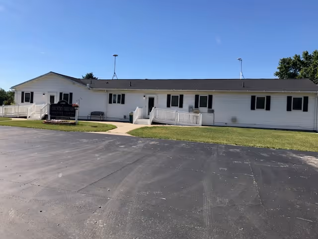 Exterior view of a single-story white building with a black roof under a clear blue sky. The building has multiple windows with black shutters and two wheelchair ramps leading to separate entrances. A sign in front reads 'Country Friends Assisted Living'. There is a paved parking area in the foreground and a grassy lawn surrounding the building.