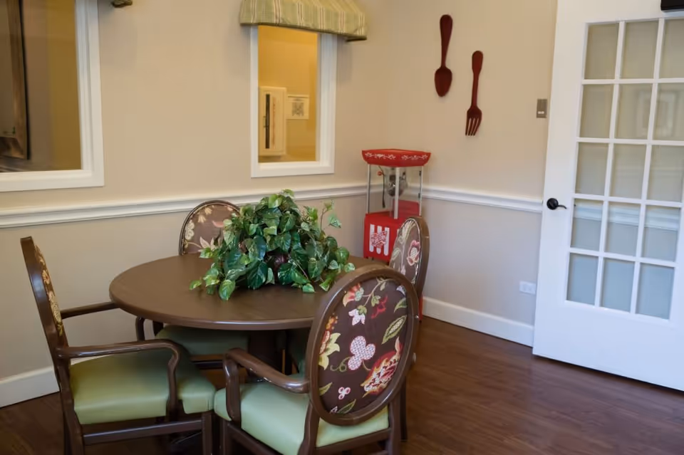 Small dining area with a round table, four upholstered chairs and a leafy centerpiece, a popcorn machine, and decorative oversized utensils on the wall.