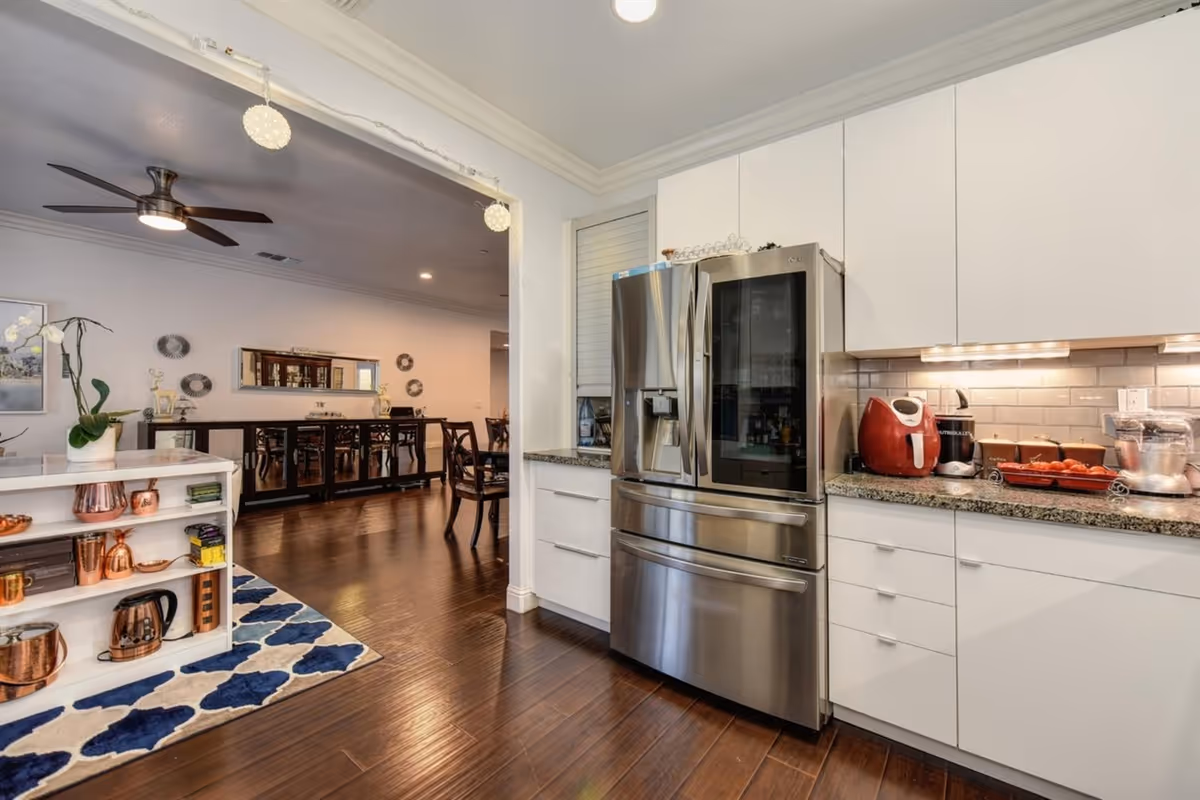 Modern kitchen area with white cabinets, a stainless steel refrigerator, and various kitchen appliances on the countertop. The kitchen opens into a dining area with a wooden floor, a ceiling fan, and a dining table with chairs. There is a small shelving unit with copper kitchenware and a blue and white patterned rug on the floor.