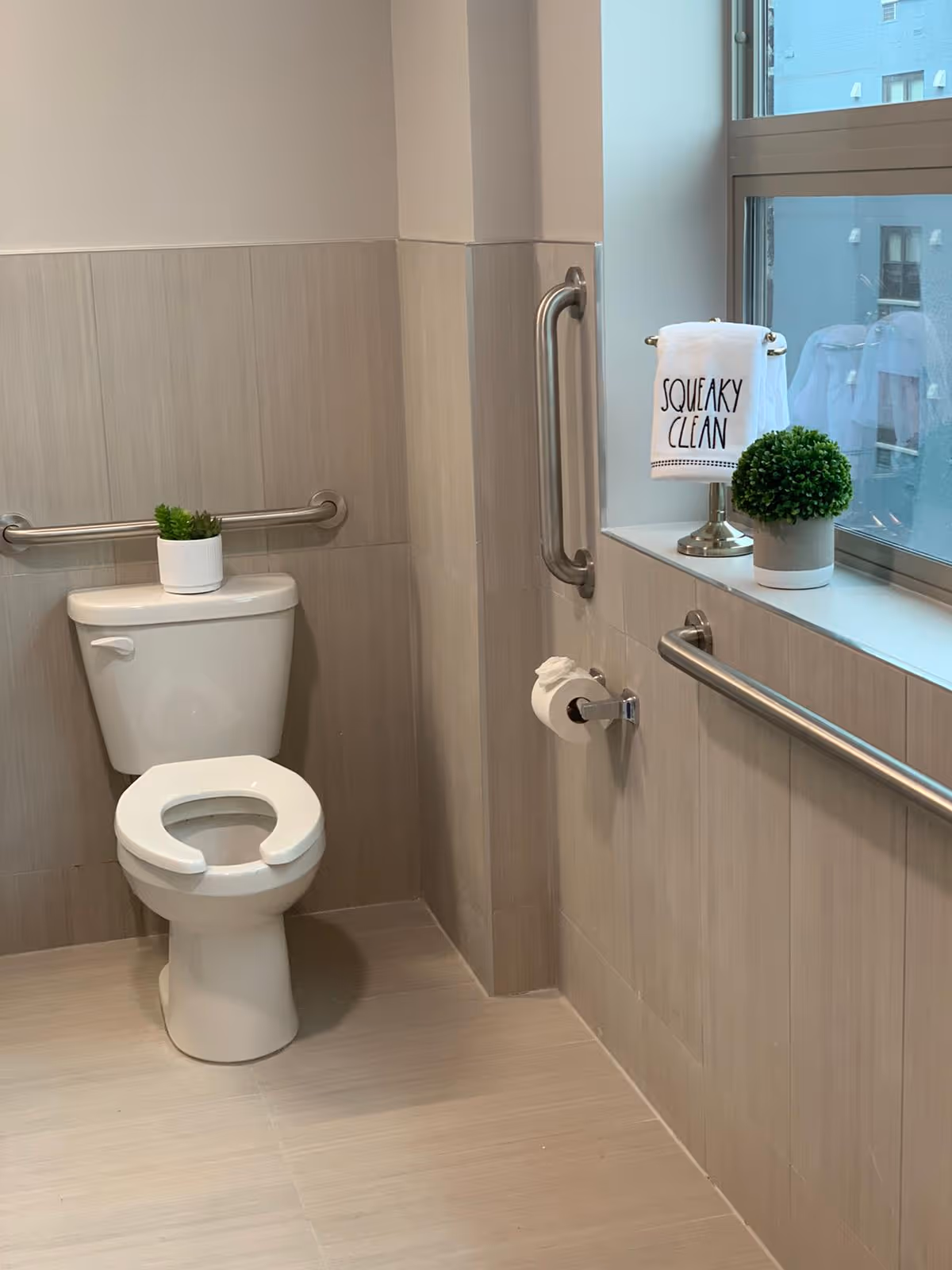 A clean and accessible bathroom with a white toilet featuring an open-front seat. There are stainless steel grab bars on the wall behind and beside the toilet. A small potted plant sits on top of the toilet tank. Next to the toilet, a toilet paper holder is mounted on the wall. A window with a ledge holds a small green potted plant and a towel on a stand that reads 'SQUEAKY CLEAN'. The walls and floor are tiled in light beige tones.