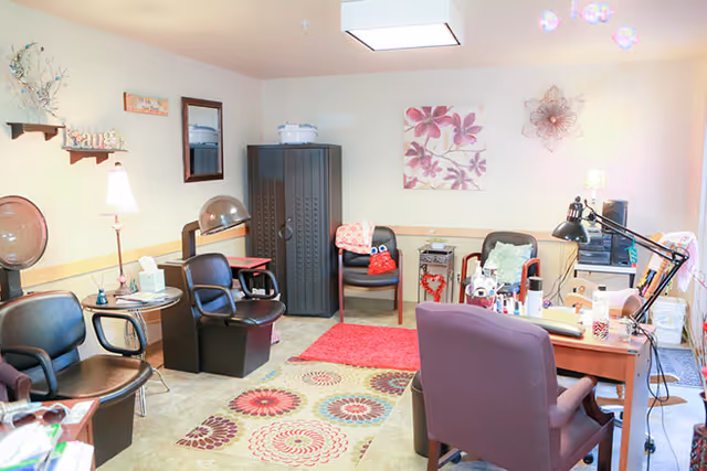 Bright salon-style interior with multiple styling chairs, hooded hair dryers, a desk, and floral wall decor.