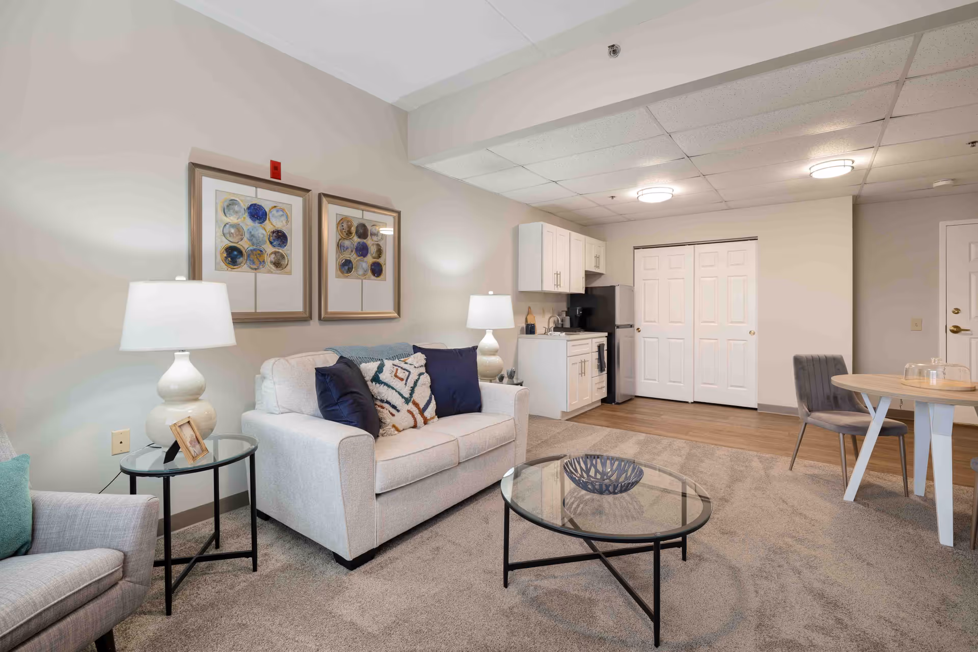 A cozy senior living community interior featuring a small living area with a beige sofa adorned with decorative pillows, a glass coffee table, and a side table with a lamp and picture frame. The background shows a compact kitchenette with white cabinets, a black refrigerator, and a small dining table with two chairs. The room has neutral-colored walls and carpeted flooring.
