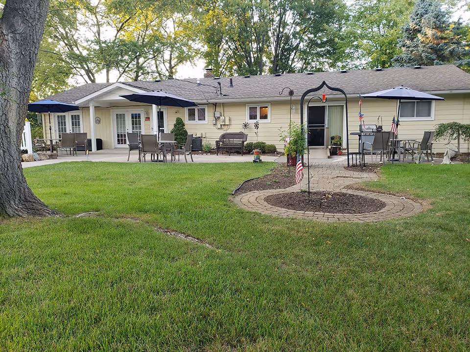 Outdoor patio area of a single-story building with beige siding, featuring multiple seating arrangements with tables and chairs under large umbrellas. There is a paved walkway leading to the entrance, surrounded by green grass and trees in the background. Small American flags and garden decorations are visible near the walkway.