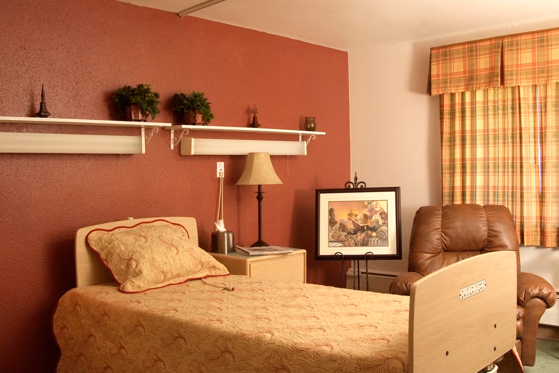 A nursing home bedroom with a single bed covered with a beige patterned bedspread and matching pillow. There is a wooden nightstand with a lamp and a tissue box next to the bed. Above the bed, two white shelves hold small potted plants and decorative items. A framed picture is displayed on a stand next to a brown leather recliner chair. The window has plaid curtains in shades of orange, yellow, and green. The walls are painted red and white.