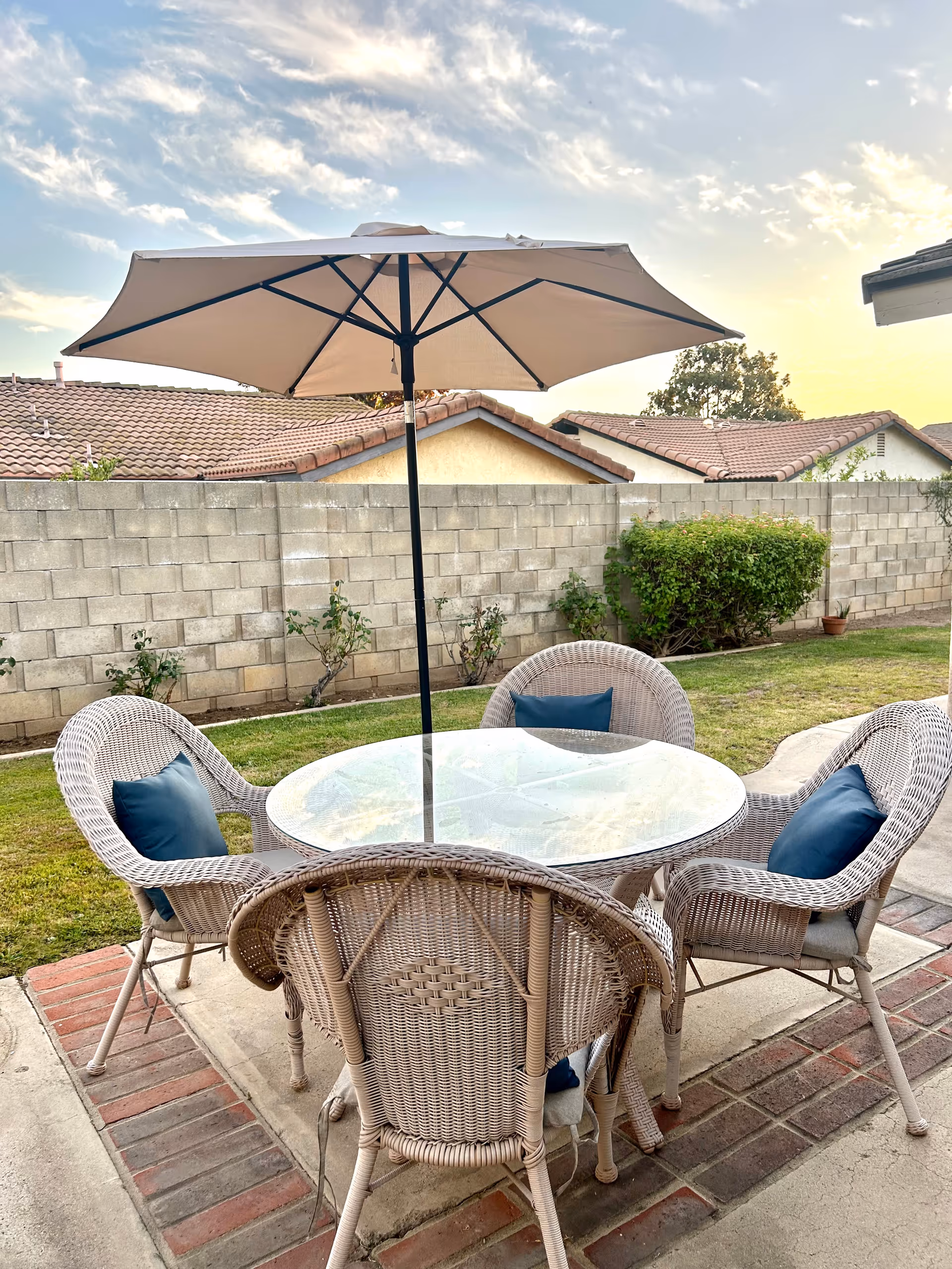 Outdoor patio area with a round glass-top table surrounded by four wicker chairs with blue cushions. A large beige umbrella is positioned in the center of the table. The patio is bordered by a brick and concrete floor, with a grass lawn and a concrete block wall in the background. Neighboring houses with tiled roofs are visible beyond the wall under a partly cloudy sky at sunset.