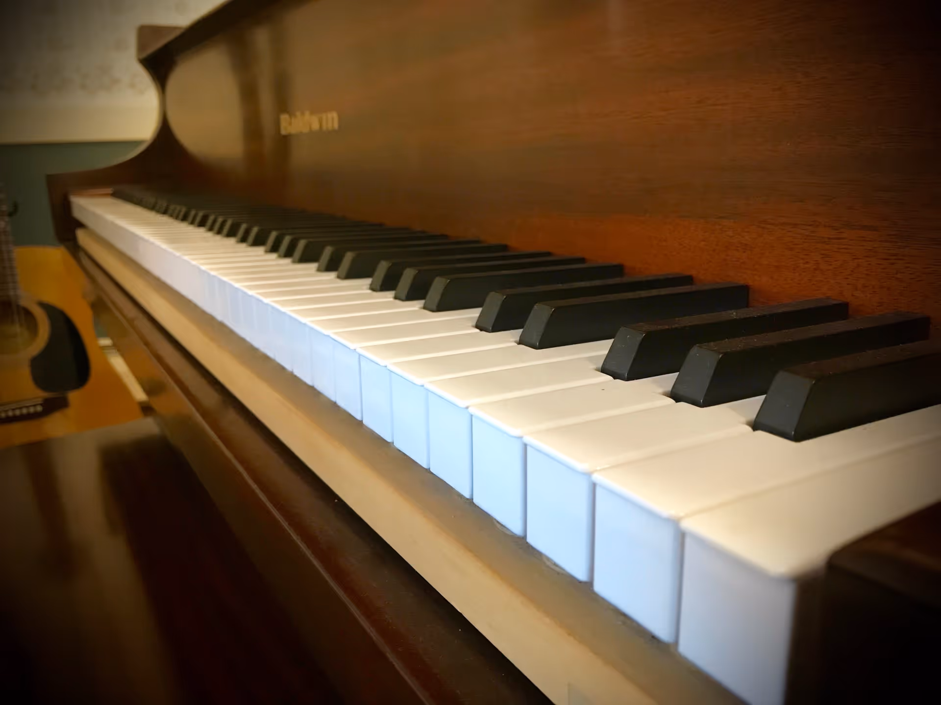 Close-up view of a Baldwin piano keyboard with white and black keys, and an acoustic guitar partially visible in the background.