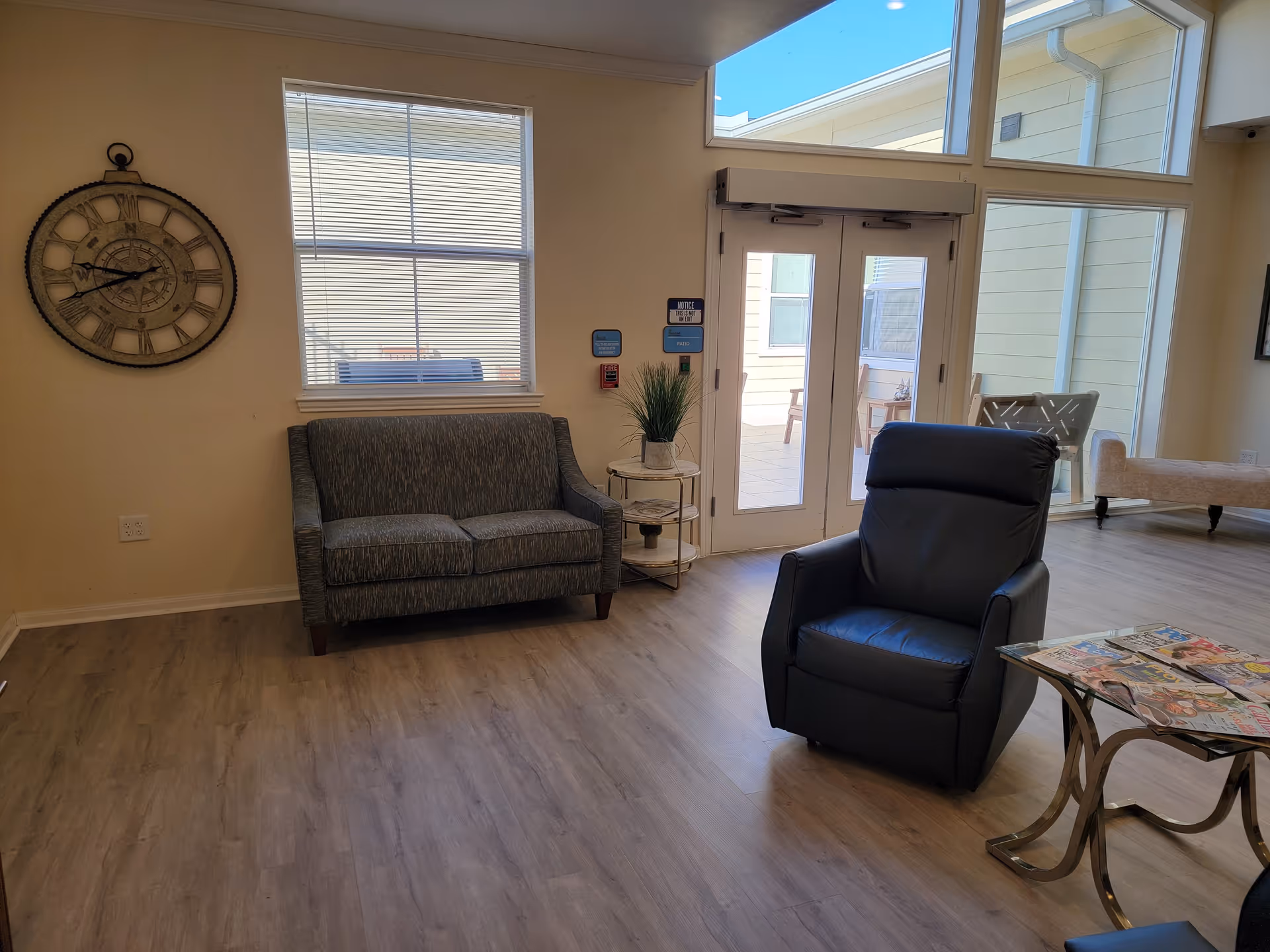 A cozy living room area with a gray loveseat, a black recliner chair, a glass-top side table with magazines, a large wall clock, and a window with blinds. There are double glass doors leading to an outdoor patio with chairs visible through the glass.