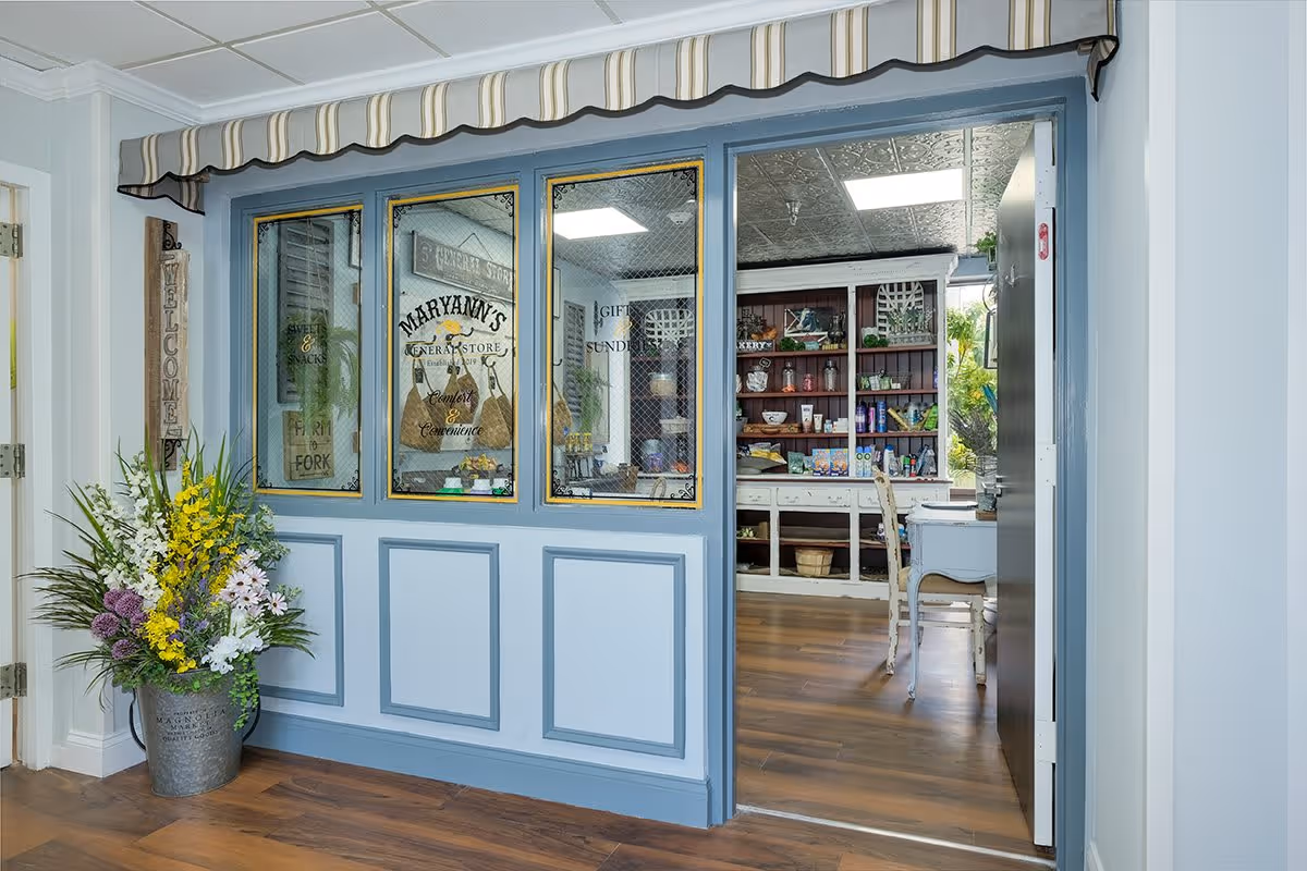 Interior view of a general store area inside a senior living facility, featuring a blue framed window with glass panes displaying the text 'Maryann's General Store Comfort & Convenience'. There is a large flower arrangement in a metal bucket to the left and shelves stocked with various items visible through the open doorway. The floor is wooden, and the ceiling has a decorative metal tile pattern.