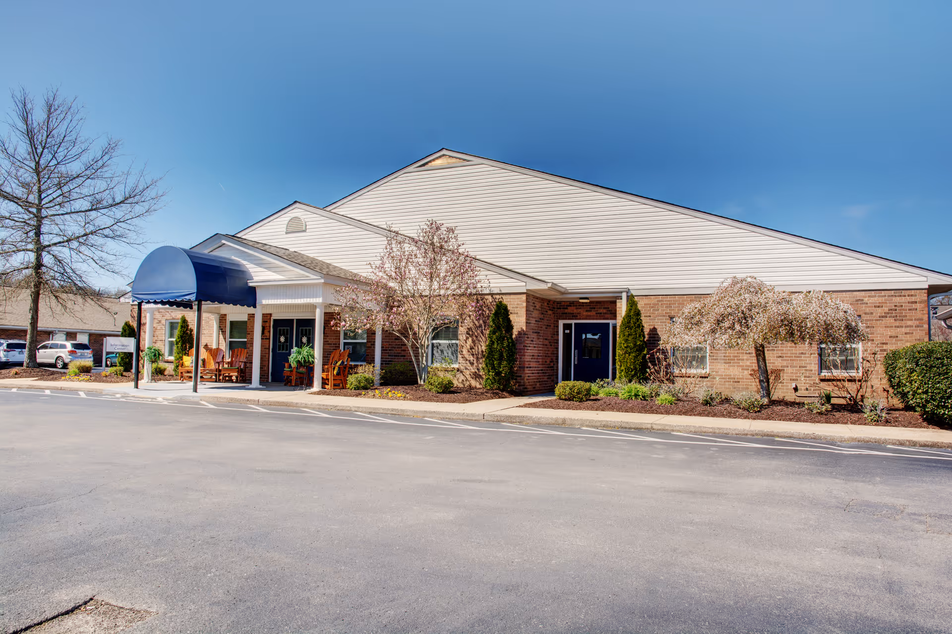 Front exterior of a single-story senior living facility with a brick facade, blue entrance awning, and landscaped shrubs and trees.