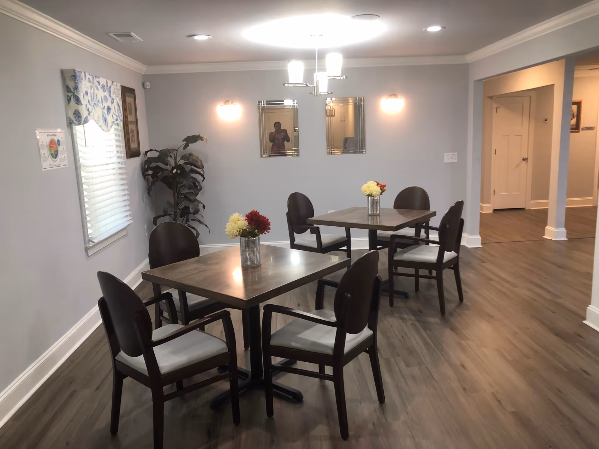 A dining area with two square wooden tables, each surrounded by four dark wooden chairs with light cushions. Each table has a small vase with colorful flowers. The room has light gray walls, wood flooring, a window with a floral valance, two decorative mirrors on the wall, and ceiling lights. There is a plant in the corner and an open doorway leading to another room.