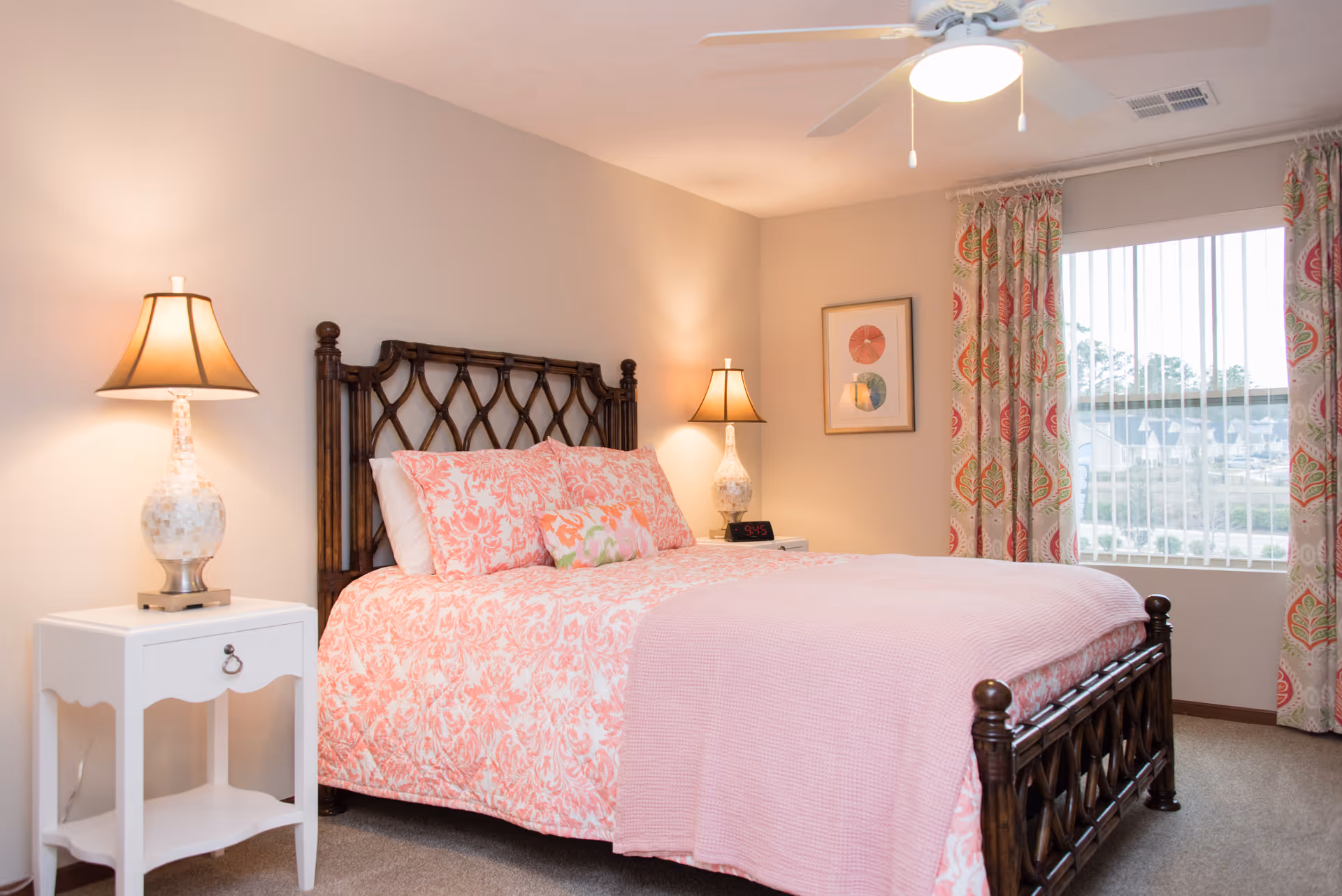 Bright bedroom with a wooden headboard, pink patterned bedding, two bedside lamps, a white nightstand, and a window with patterned curtains.