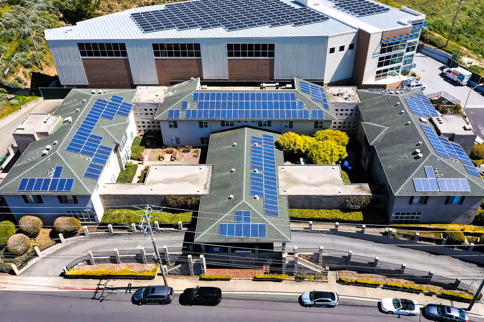 Aerial view of Mission Villa Senior Living facility showing multiple buildings with green roofs covered in solar panels. The buildings are arranged around a central courtyard with trees and bushes. A large industrial building with more solar panels is visible behind the facility. Several cars are parked along the street in front of the property.